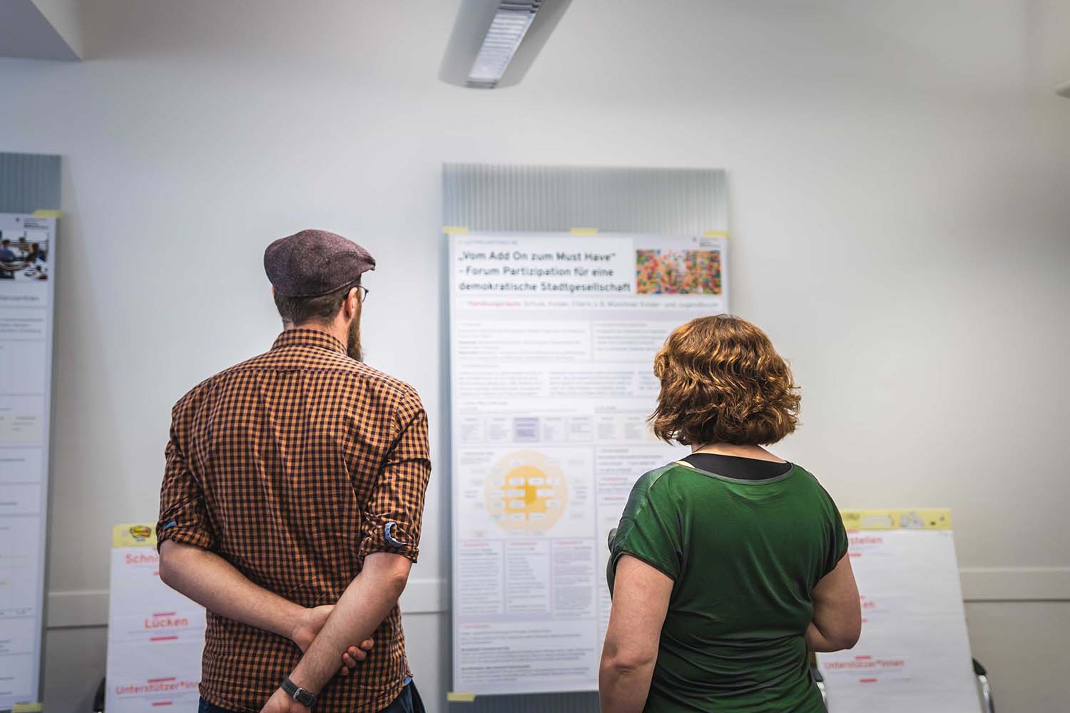 Man sieht zwei Personen von hinten, die vor einem großen Plakat stehen, das über ein Forum zur demokratischen Beteiligung informiert. Die erste Person, die ein kariertes Hemd und eine flache Mütze trägt, hat die Arme hinter dem Rücken verschränkt. Die zweite Person, mit lockigem Haar und grünem Oberteil, steht neben ihr, beide auf den Inhalt des Plakats konzentriert. Die Umgebung scheint ein moderner Innenraum zu sein, wahrscheinlich während einer Präsentation oder Informationsveranstaltung.